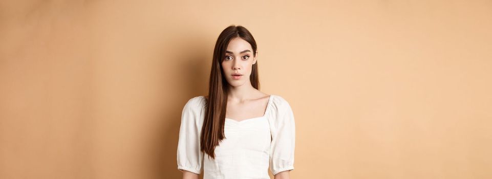 Shocked and startled young woman freeze in awe, open mouth and look at camera amazed, standing in dress on beige background.