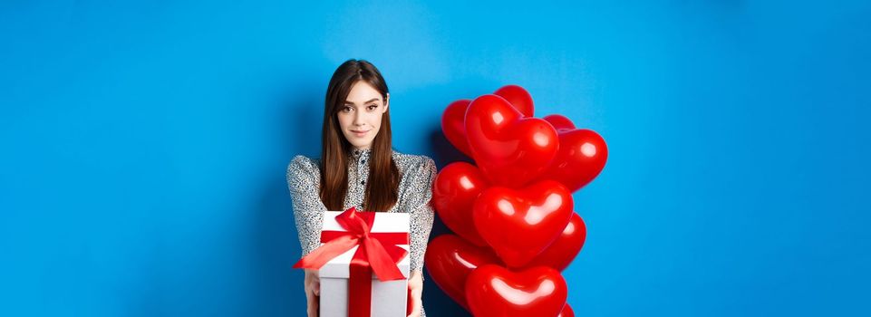 Valentines day. Romantic and cute girl stretching out hands with present, giving gift box to lover and smiling, standing near hearts balloons, blue background.