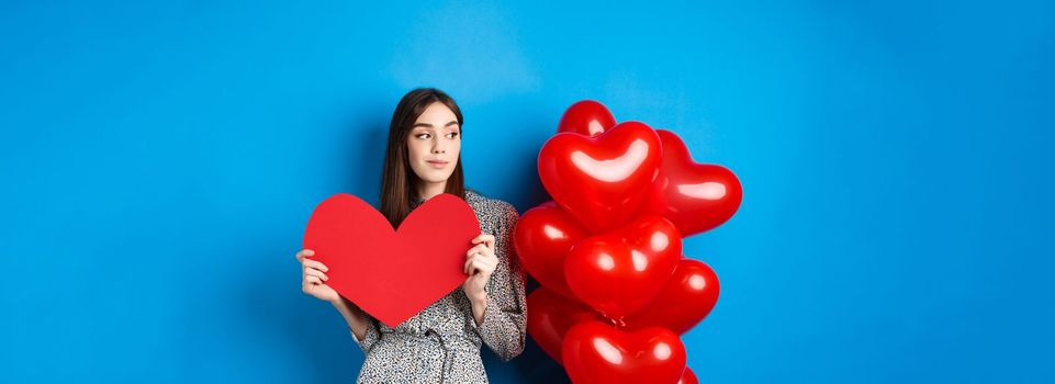 Valentines day. Romantic girl in dress standing near balloons and holding big red heart cutout, dream of something, standing on blue background.