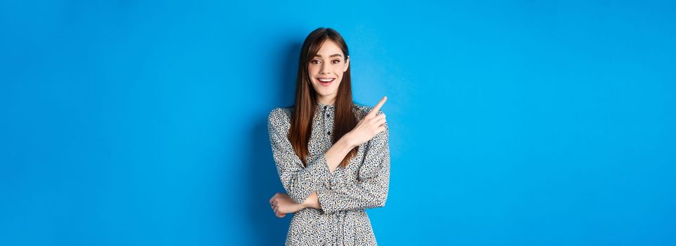 Cheerful young woman in long dress smiling, pointing finger at upper left corner, advertising product on blue background.