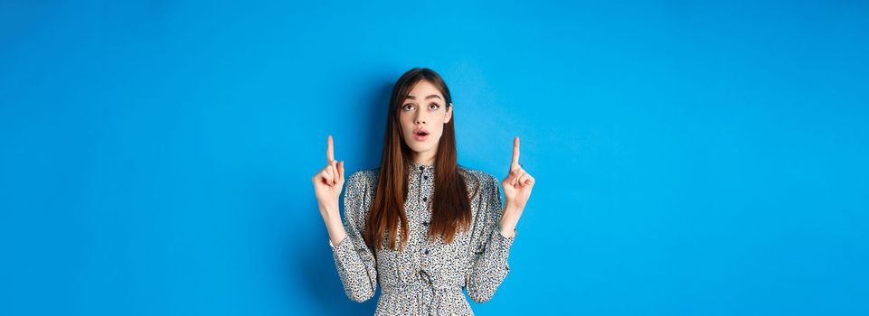 Surprised romantic girl in dress pointing, looking up with opened mouth and fascinated gaze, standing on blue background.