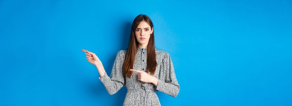 Angry frowning woman pointing fingers right and condemn person, feeling disapointed, standing in dress on blue background.