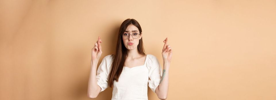 Cute hopeful girl making wish, pucker lips and begging, say please, standing on beige background.