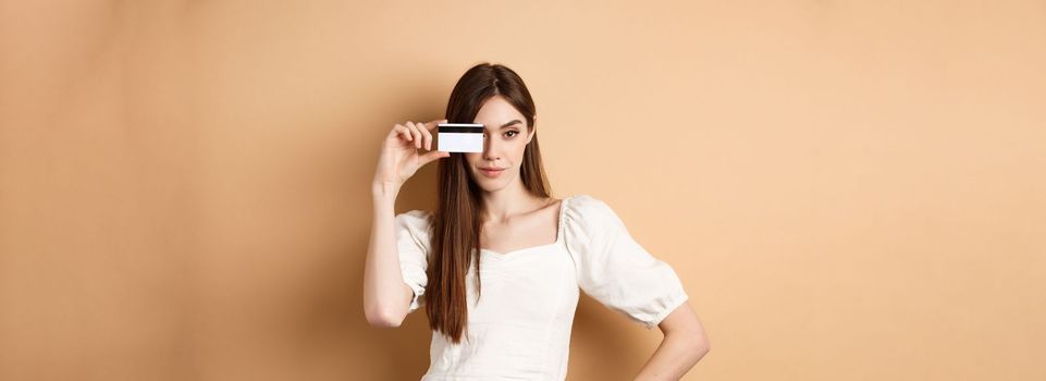 Sassy young woman showing plastic credit card on face and looking determined, going on shopping, standing against beige background.