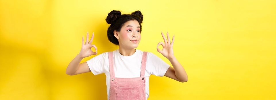 Romantic and beautiful asian girl looking at logo left, showing okay signs in approval, standing on yellow background.