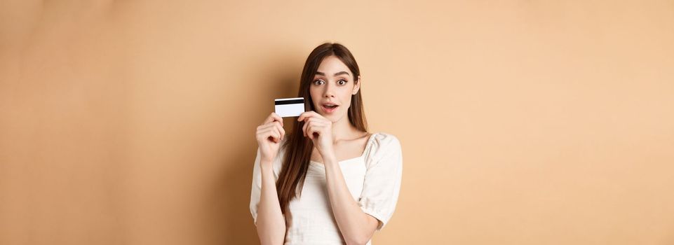Excited young woman showing plastic credit card and look amazed at camera, standing on beige background.