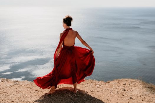 Side view a Young beautiful sensual woman in a red long dress posing on a rock high above the sea during sunrise. Girl on the nature on blue sky background. Fashion photo.