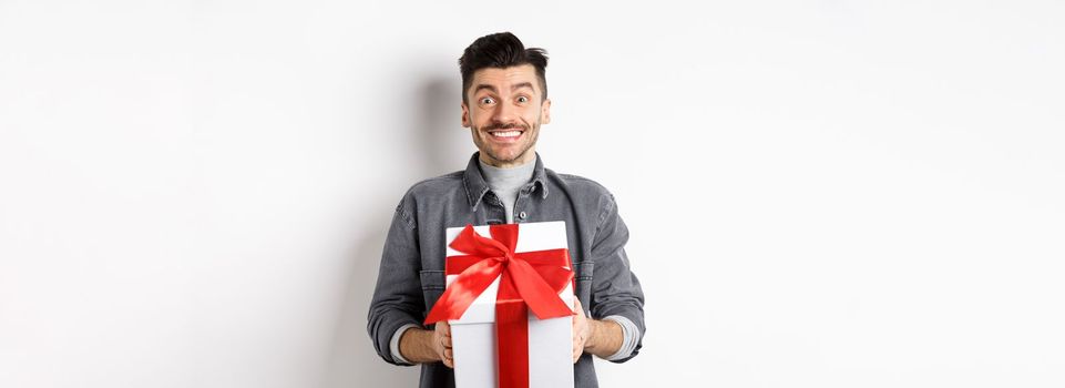 Cheerful caucasian guy holding surprise gift, receive present on holiday and smiling thankful, looking grateful at camera, celebrating Valentines day, white background.