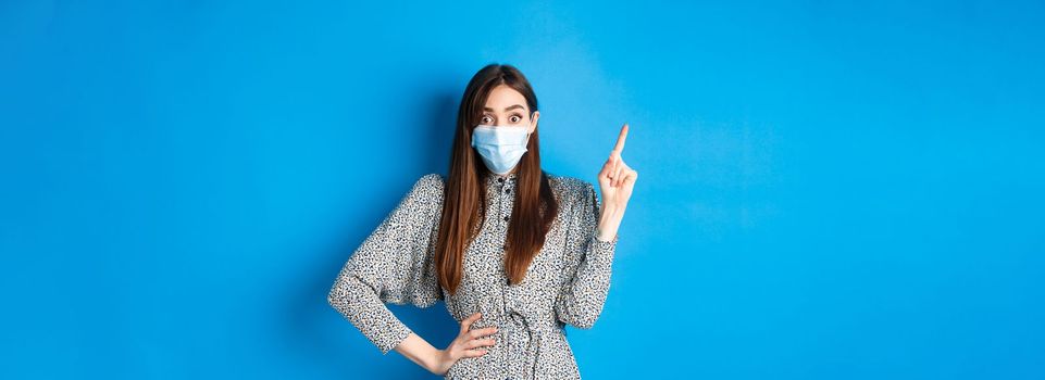Healthy people and covid-19 pandemic concept. Excited woman in medical mask pointing at upper left corner, standing on blue background.