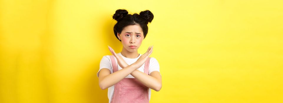 Sad girl begging to stop, frowning uspet and showing cross sign, say no, standing gloomy on yellow background.