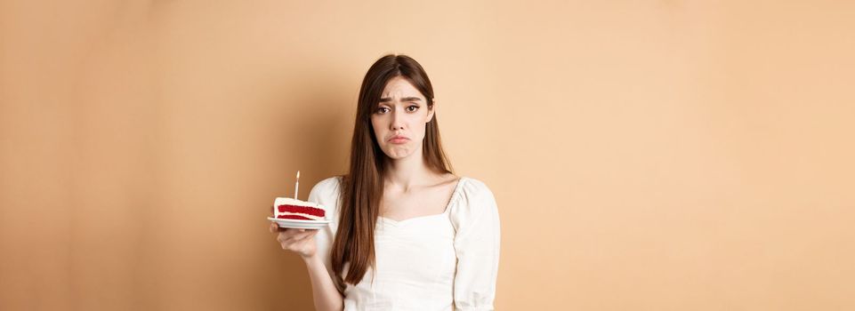 Sad birthday girl holding cake with candle and look upset, feeling lonely on her bday, standing alone on beige background.