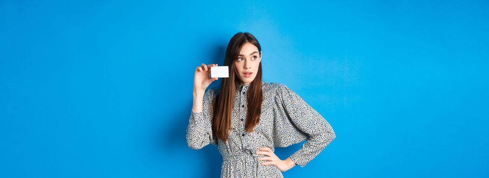 Shopping. Thoughtful female shopper looking left at logo, showing plastic credit card, standing on blue background.