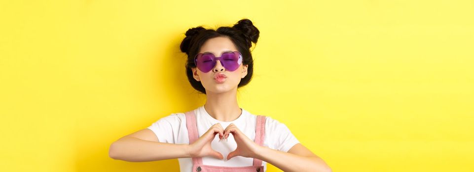 Summer and fashion concept. Cute glam girl with sunglasses and hairbuns, showing heart sign and pucker lips for kiss, I love you gesture, yellow background.