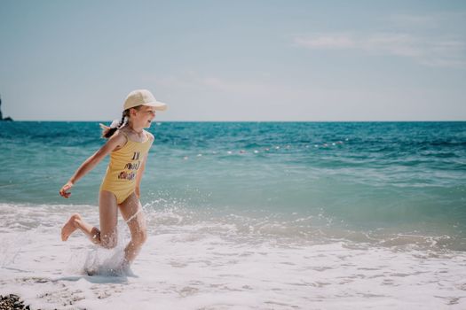 Cute little girl running along the seashore against a clear blue sea and rejoices in the rays of the summer sun. Beautiful girl in yellow swimsuit running and having fun on tropical beach