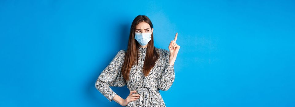 People, covid and quarantine concept. Suspicious frowning girl in medical mask, pointing and looking aside with hesitant face, standing on blue background.