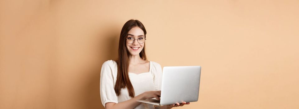 Smiling girl student working on laptop, wearing glasses and looking happy, using computer while standing against beige background.