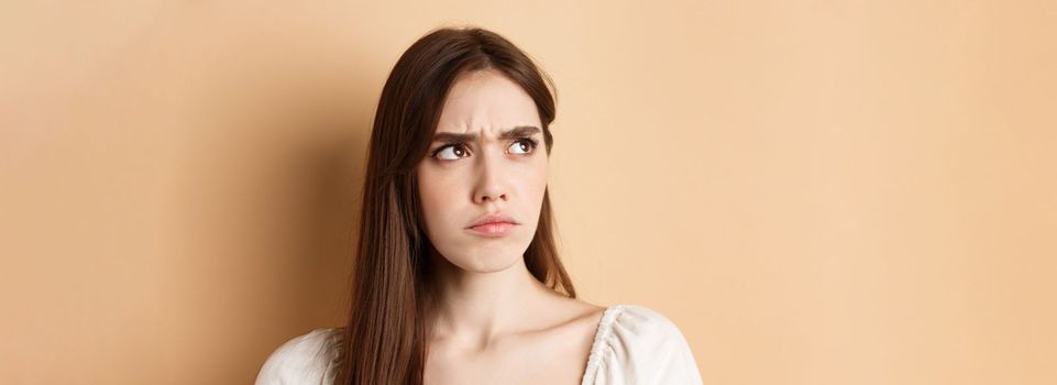 Close up of angry cute girl frowning, looking aside displeased, stare at bad thing, standing on beige background.