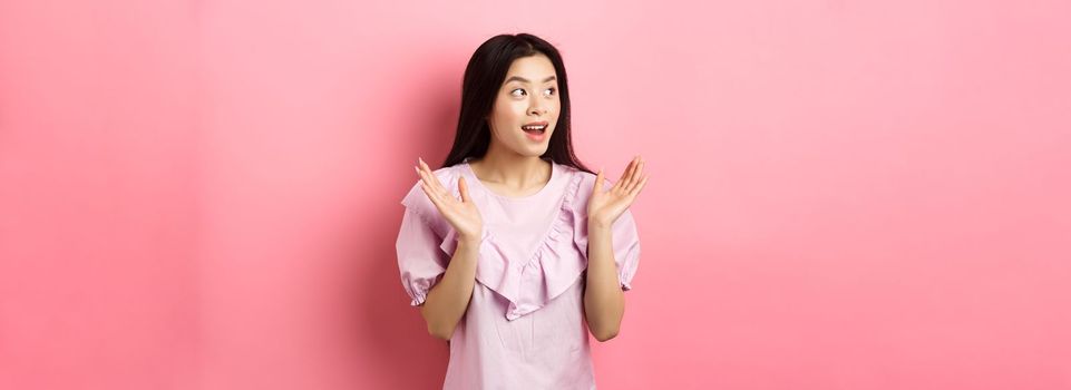 Excited asian girl clap hands and looking left, watching performance and applause, standing in dress on pink background.