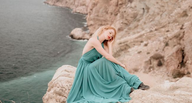 Side view a Young beautiful sensual woman in a mint long dress posing on a volcanic rock high above the sea during sunset. Girl on the nature on overcast sky background. Fashion photo