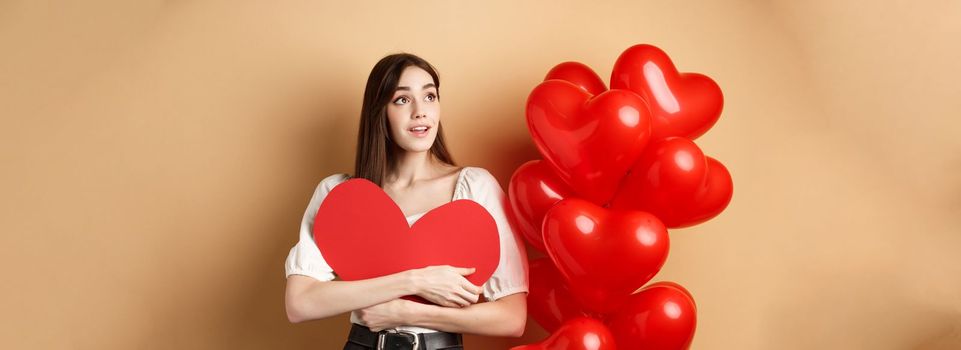 Romantic girl hugging Valentine day big red heart cutout, standing near romance balloons and looking left at logo, gazing dreamy at promo, beige background.