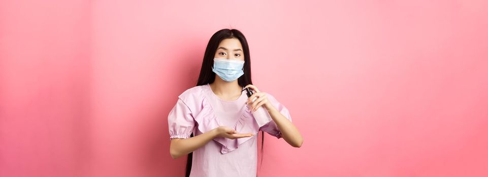 Healthy people and covid-19 pandemic concept. Cute asian woman in medical mask clean hands with antiseptic, using sanitizer, standing against pink background.