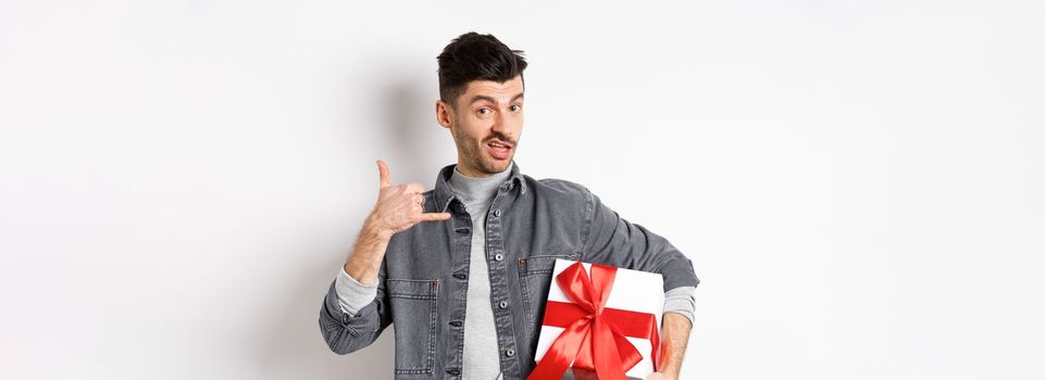 Macho guy with gift box show call me phone gesture, flirting with you, standing boastful on white background. Relationship and Valentines day concept.