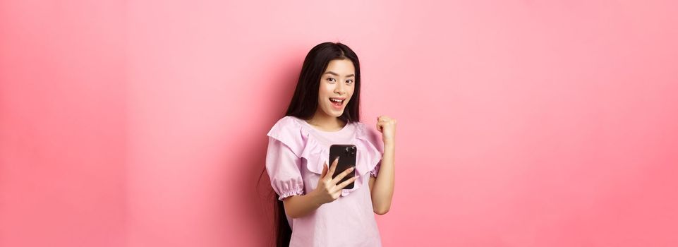 Online shopping. Satisfied asian girl winning on mobile phone, say yes and make fist pump, holding smartphone, standing against pink background.