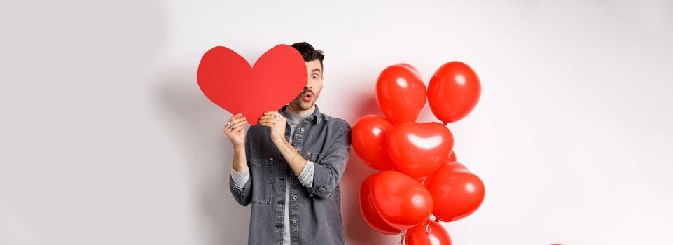 Cute young man hiding face behind big Valentine heart card, secret admirer looking at camera with amazement, saying wow, standing near romantic balloons on white background.