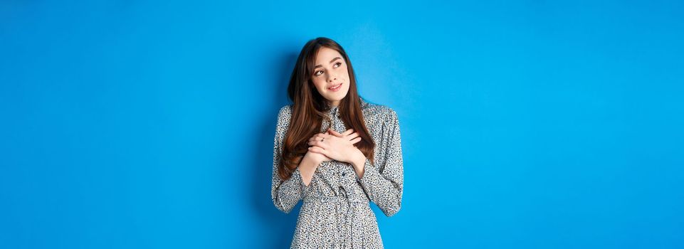 Romantic dreamy girl in dress looking at upper left corner, holding hands on heart and daydreaming, imaging something cute, standing on blue background.