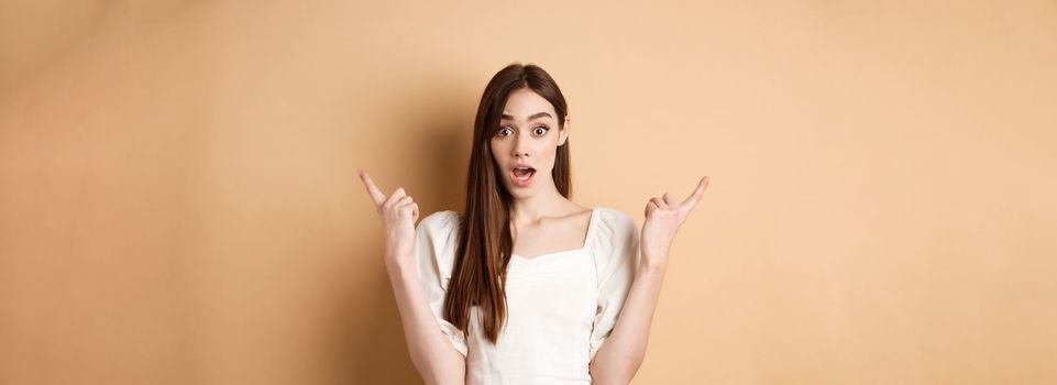 Surprised young girl pointing sideways at left and right, gasping and saying wow amazed, showing two cool choices, beige background.