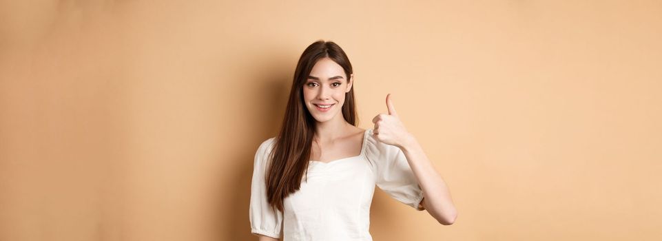 Good job. Smiling candid woman in blouse showing thumb up, praise nice choice, recommending product, standing satisfied on beige background.