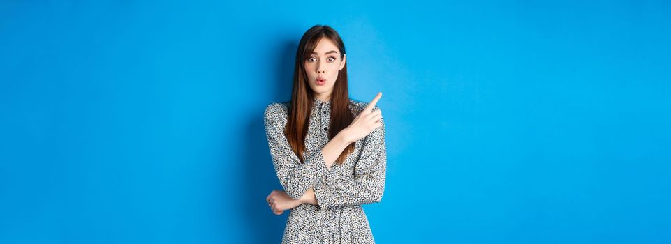 Excited young woman say wow, looking impressed and pointing left at logo, showing awesome news, standing cute dress on blue background.