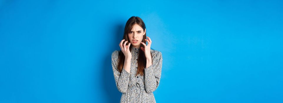 Angry jealous girl clench hands in fists, grimacing furious, standing mad and outraged on blue background.