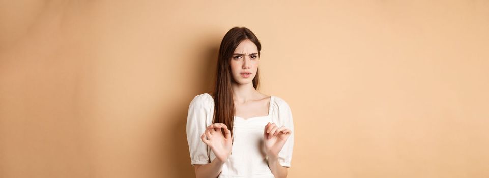 Stay away from me. Disgusted young woman frowning and looking with aversion at something stinky, rejecting disgusting thing, standing on beige background.