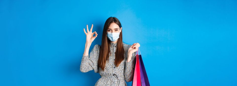 Covid-19, pandemic and lifestyle concept. Attractive woman wear medical mask on shopping, show okay and hold bags with purchases, blue background.