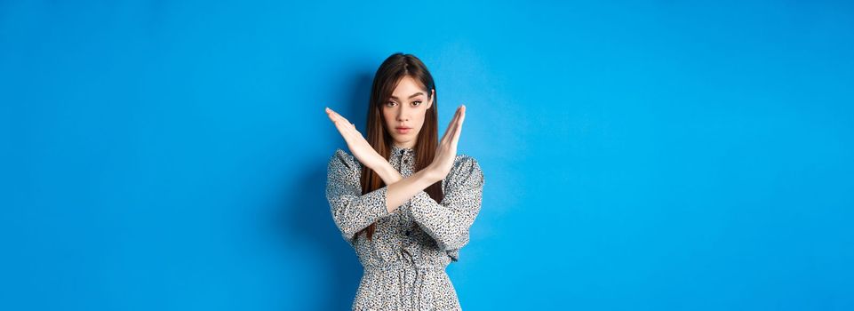 Serious caucasian woman in dress tell no, show cross stop gesture and looking confident, making warning sign, prohibit something bad, standing on blue background.