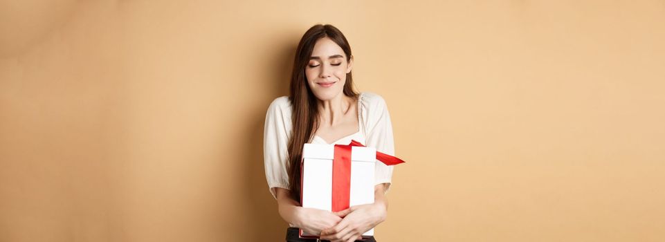 Happy valentines day. Dreamy woman hugging her surprise gift, close eyes and smile, standing on beige background.