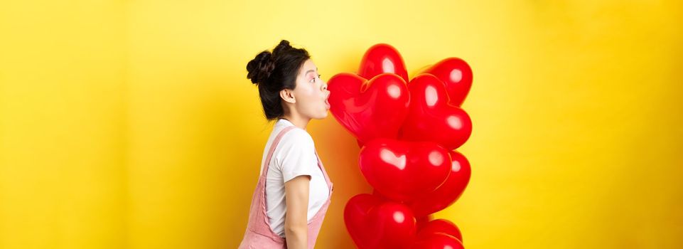 Valentines day and relationship concept. Profile of young asian woman scream of surprise, say wow and looking left amazed, standing near red balloons, yellow background.