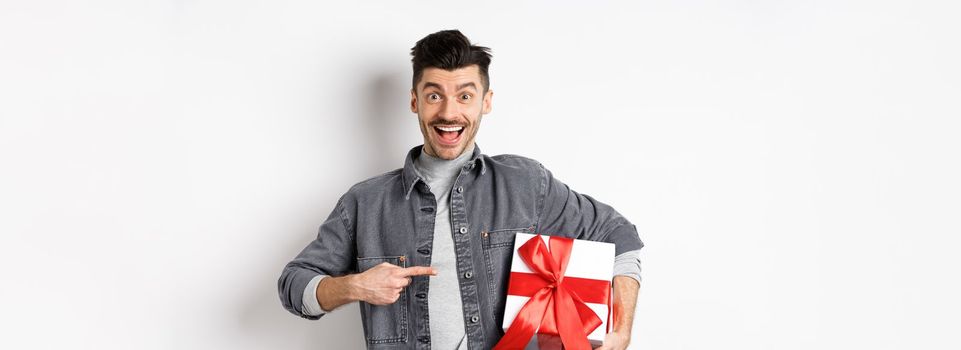 Happy Valentines day. Excited caucasian guy pointing at gift box from lover, smiling and looking amazed, buying presents on romantic date, white background.