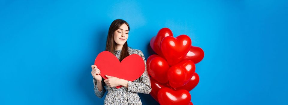 Valentines day. Romantic smiling woman hugging big red heart cutout and dreaming of love, standing in dress on blue background.