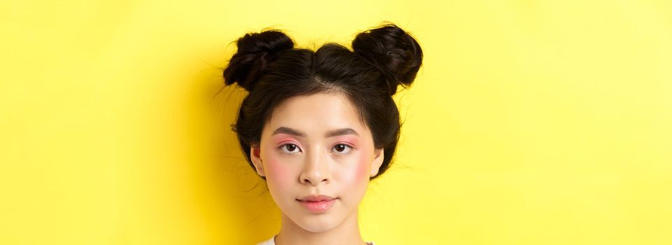 Head portrait of stylish asian girl with bright makeup and hairbuns, looking at camera, standing on yellow background.