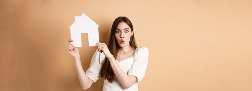 Real estate. Excited woman showing paper house cutout and saying wow with amazed face, buying property, standing on beige background.