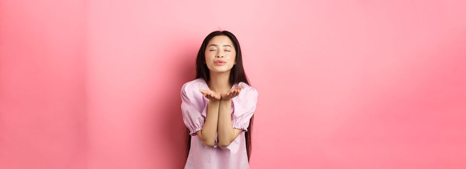 Cute and romantic asian girl blowing air kiss at camera, close eyes and smile tenderly, standing in dress against pink background.