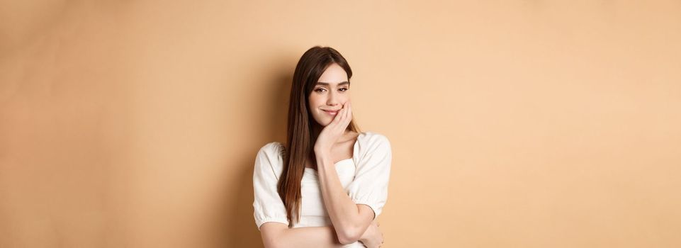 Romantic young woman touching face and smiling at camera with pensive look, standing dreamy on beige background.