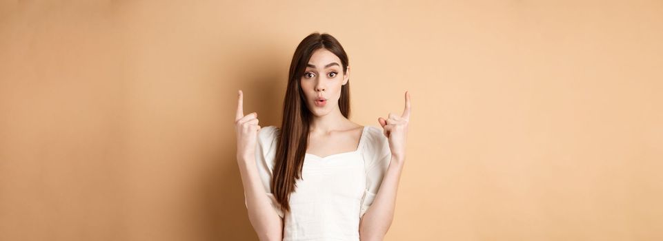 Wow look. Intrigued young woman showing interesting offer, pointing fingers up and look excited, standing on beige background.