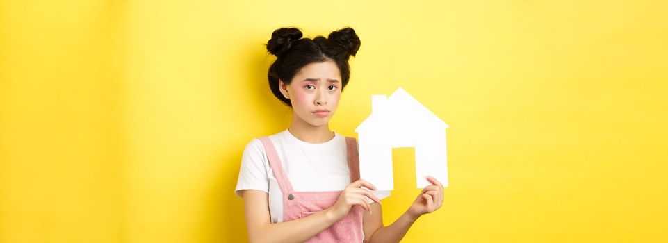 Real estate and family concept. Sad cute asian girl with bright makeup, frowning and feel upset, showing paper house cutout, standing on yellow background.