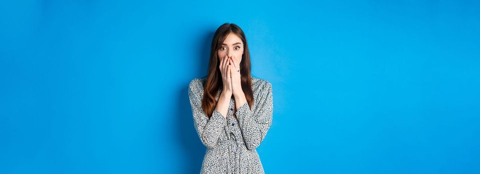 Shocked european girl gasping and covering mouth with hands, witness something and look startled, standing worried on blue background.