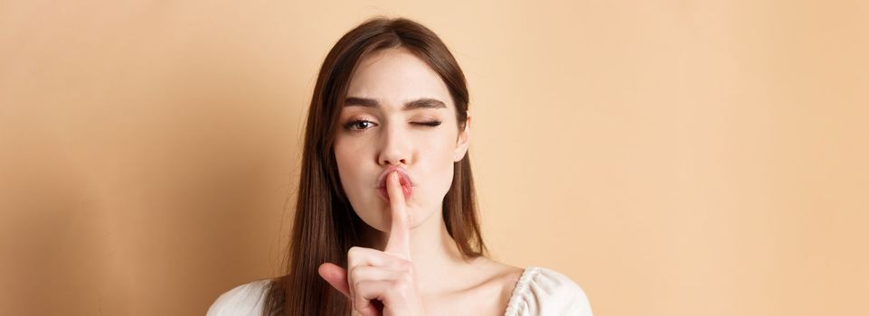 Close up of attractive woman shushing at camera, hiding secret, making hush sign and winking, show taboo sign, standing on beige background.