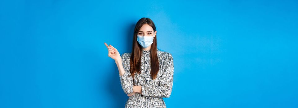 Covid-19, social distancing and healthcare concept. Pretty lady in medical mask showing way, pointing right and smiling, advertising on blue background.