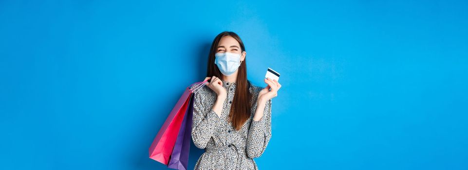 Covid-19, pandemic and lifestyle concept. Happy girl wear face mask on shopping, showing plastic credit card and laughing carefree, buying gifts, standing on blue background.